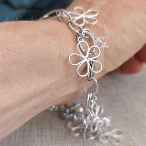May include: A silver-toned charm bracelet featuring multiple open-work flower motifs connected by circular links. The bracelet includes small, clear, faceted crystal beads interspersed among the metallic floral shapes, creating a layered design.