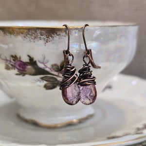Puede incluir: Pendientes hechos a mano con piedras moradas en forma de lágrima envueltas en alambre de cobre. Los pendientes cuelgan de ganchos de cobre. Fondo: taza de té vintage con diseños florales.