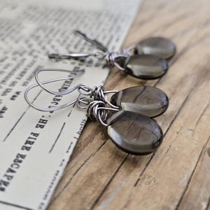 May include: A pair of smoky quartz earrings with silver-colored wire wrapping. The teardrop-shaped stones are a translucent gray color. The earrings are displayed on a vintage newspaper background and a wooden surface.