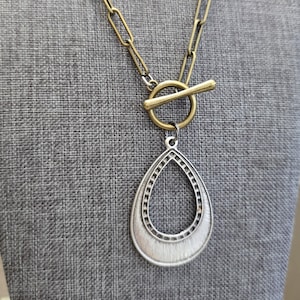 Op de afbeelding: Een gouden ketting met een toggle-sluiting en een zilveren traanvormige hanger. De hanger heeft een getextureerd ontwerp en een ronde opening in het midden.