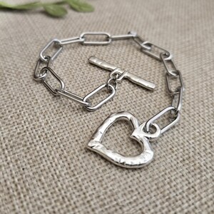 Pu&ograve; includere: Un braccialetto a catena color argento con un ciondolo a forma di cuore. Il braccialetto presenta una chiusura a levetta e maglie rettangolari. Il ciondolo a cuore ha una superficie strutturata.