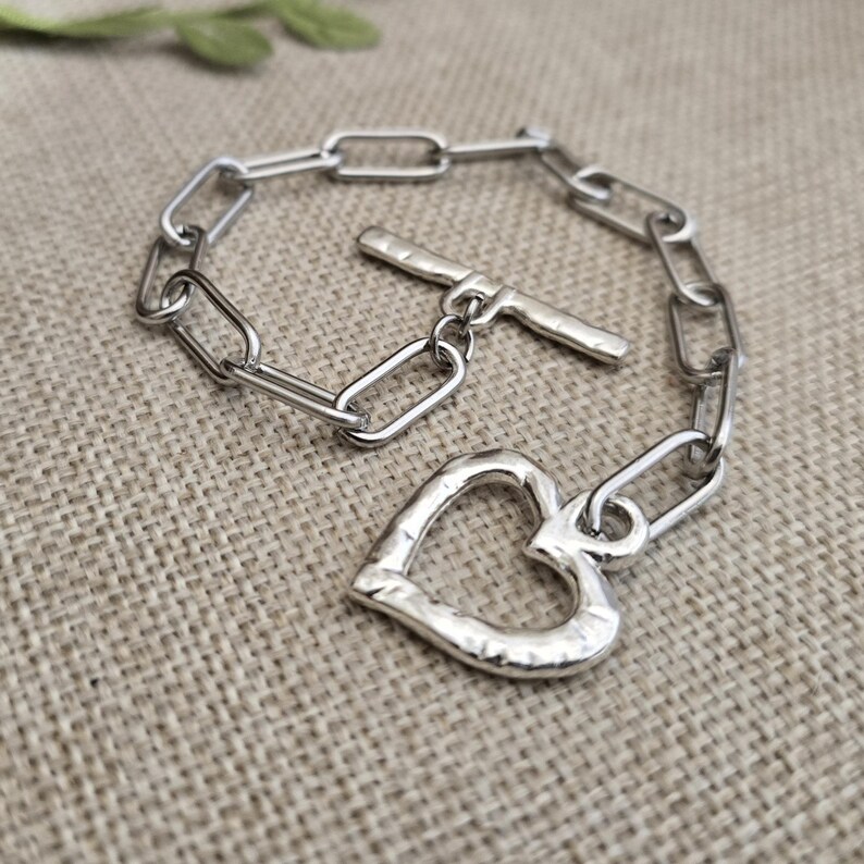 Pu&ograve; includere: Un braccialetto a catena color argento con un ciondolo a forma di cuore. Il braccialetto presenta una chiusura a levetta e maglie rettangolari. Il ciondolo a cuore ha una superficie strutturata.