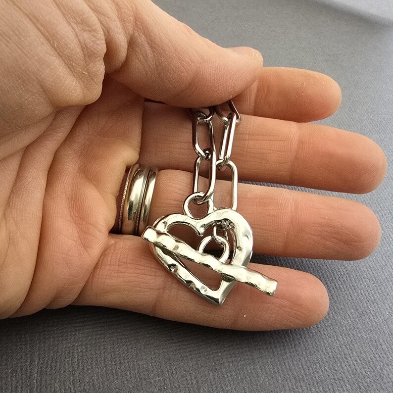 Pu&ograve; includere: Una collana con pendente a forma di cuore argentato con chiusura a toggle. Il cuore ha una superficie strutturata e una barra che lo attraversa. La collana &egrave; tenuta in una mano, con un anello d'argento al dito.