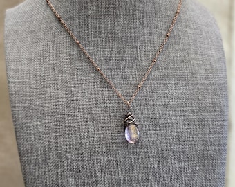 May include: A copper-colored necklace with a small, teardrop-shaped pendant. The pendant is a light purple color and is wrapped in copper wire. The necklace chain is delicate and has small, round beads spaced along its length.