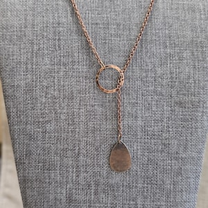 May include: Copper-colored necklace with a hammered circle pendant and a teardrop-shaped charm. The necklace hangs from a delicate chain, creating a Y-shaped design. The jewelry is displayed on a gray fabric background.