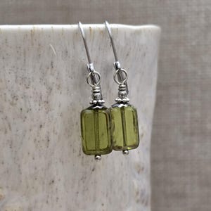 Puede incluir: Un par de pendientes colgantes con cuentas rectangulares de color verde oliva. Los pendientes tienen ganchos plateados y detalles decorativos plateados. Las cuentas son translúcidas. Fondo neutro.