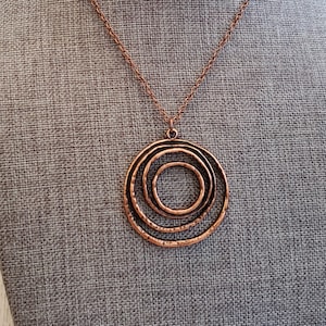 May include: A copper necklace with a large, circular pendant featuring three concentric circles. The pendant has a hammered texture.