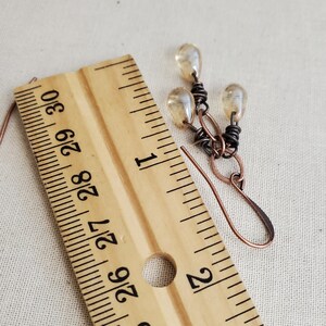 K&ouml;nnte beinhalten: Ohrringe aus Kupferdraht mit drei kleinen, klaren Glasperlen, die am unteren Ende h&auml;ngen. Die Ohrringe sind etwa 2,5 cm lang und werden neben einem Lineal gezeigt.