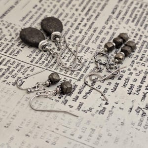 May include: A collection of handcrafted earrings featuring dark gray, textured stones and silver-toned metal components. The earrings showcase various designs, including teardrop shapes and geometric beads, set against a vintage newspaper background.