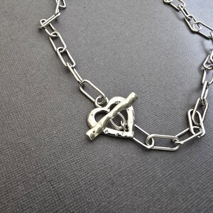 Pu&ograve; includere: Collana color argento con chiusura a forma di cuore. Il design a catena presenta un ciondolo a forma di cuore e una chiusura a barra. Il gioiello &egrave; esposto su una superficie grigia.