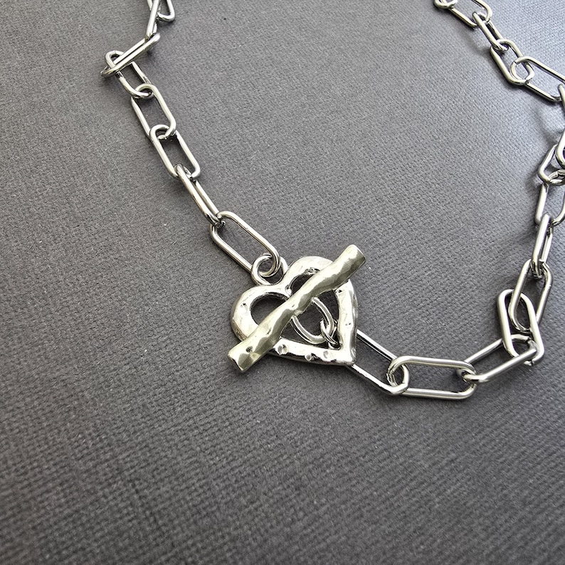 Pu&ograve; includere: Collana color argento con chiusura a forma di cuore. Il design a catena presenta un ciondolo a forma di cuore e una chiusura a barra. Il gioiello &egrave; esposto su una superficie grigia.