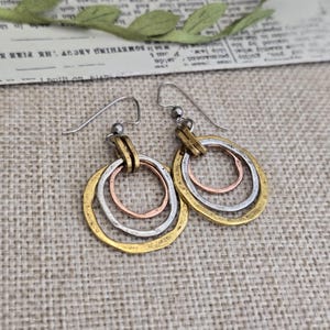 May include: A pair of dangle earrings featuring three interlocking circular rings in gold, silver, and copper tones. Each earring hangs from a simple silver-toned hook wire. The metal rings have a textured, hammered finish and are connected by a small metal loop.