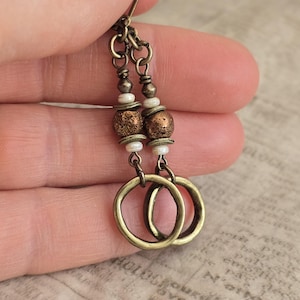 May include: A pair of antique brass earrings. Each earring features a chain with small beads, textured copper-colored beads, and white beads, leading to a pair of interlocking brass hoops. The earrings are designed for pierced ears.