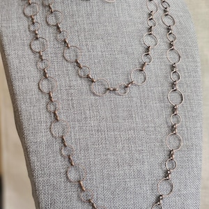 Puede incluir: Un conjunto de joyas de color cobre que incluye un collar largo, un collar más corto y un par de pendientes. Las piezas están hechas de círculos interconectados, creando un diseño similar a una cadena. El conjunto se exhibe sobre una superficie de tela gris.