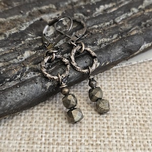May include: A pair of drop earrings featuring dark metallic lever-back hooks attached to textured circular rings. Each earring suspends two faceted, cube-shaped metallic beads in a gunmetal gray tone, creating a dangling, multi-layered design.