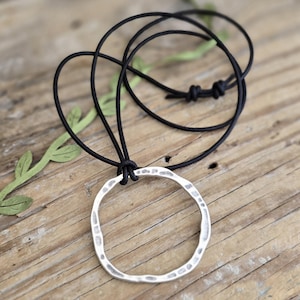 Puede incluir: Un collar con un colgante circular de plata con textura martillada, que cuelga de un cordón de cuero negro.