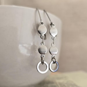 May include: A pair of silver-toned dangle earrings. Each earring features three circular discs connected by small chains, culminating in a small open circle at the bottom. The earrings hang from a simple hook.
