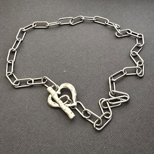 Pu&ograve; includere: Una collana color argento con un design a catena. La collana ha una chiusura a forma di cuore. Le maglie sono di forma ovale e collegate. La collana &egrave; esposta su una superficie grigio scuro.