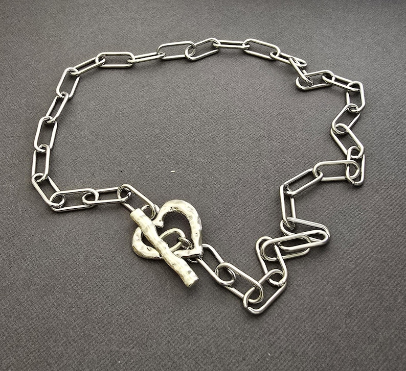 Pu&ograve; includere: Una collana color argento con un design a catena. La collana ha una chiusura a forma di cuore. Le maglie sono di forma ovale e collegate. La collana &egrave; esposta su una superficie grigio scuro.