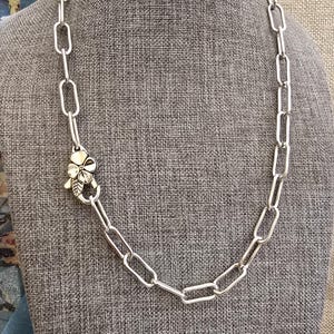 Puede incluir: Un collar de cadena de tono plateado con eslabones rectangulares alargados y un cierre prominente en forma de flor. El cierre está colocado en el lateral de la cadena y muestra un diseño floral de cuatro pétalos con un pequeño detalle de hoja.