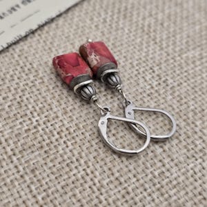 May include: A pair of dangle earrings featuring rectangular red and tan stone beads with silver-colored metal accents. The earrings have lever-back closures and are displayed on a textured, neutral-toned surface.