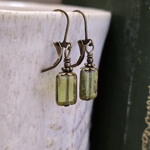 May include: A pair of antique-style earrings with olive green rectangular glass beads. The earrings have bronze-colored ear wires and decorative caps. The earrings are displayed against a blurred background of a white ceramic cup and a green book.