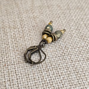 May include: A pair of handcrafted earrings featuring a bronze-colored clasp and decorative beads. The earrings have a mix of olive green and tan-colored beads, with a textured, antique finish. The earrings are designed for pierced ears.