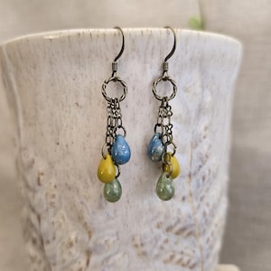 Puede incluir: Un par de pendientes colgantes de estética vintage. Cada pendiente presenta un gancho y una cadena plateados, con un grupo de cuentas coloridas en forma de lágrima en azul, amarillo y verde. Los pendientes se muestran sobre un fondo blanco.