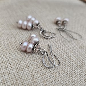 May include: Three pairs of light pink pearl earrings with silver-colored hooks and decorative accents. The earrings feature a cluster of pearls and small beads, with a simple, elegant design. The background is a neutral, textured fabric.