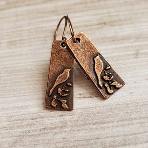 May include: A pair of copper earrings with a bird and branch design. The earrings are a rectangular shape with a rustic finish.