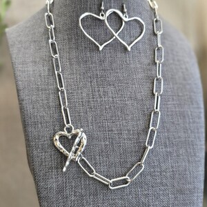 Pu&ograve; includere: Un set di gioielli color argento con collana e orecchini. La collana ha una grande chiusura a forma di cuore e una catena a maglie rettangolari. Gli orecchini sono a forma di cuore. I gioielli sono esposti su un manichino grigio.