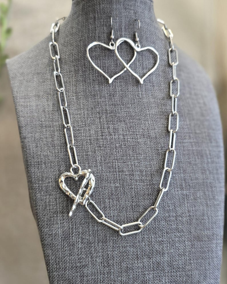 Pu&ograve; includere: Un set di gioielli color argento con collana e orecchini. La collana ha una grande chiusura a forma di cuore e una catena a maglie rettangolari. Gli orecchini sono a forma di cuore. I gioielli sono esposti su un manichino grigio.