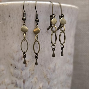 May include: Three pairs of antique brass earrings. Each earring features a hook, a small bead, a flat oval disc, an elongated oval link, and a teardrop-shaped charm. The earrings are displayed against a textured, off-white background.