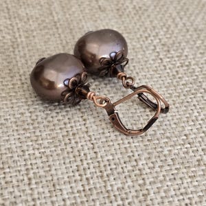 May include: A pair of drop earrings featuring round, dark brown, lustrous beads. Each bead is accented with a copper-toned metal flower-shaped cap and a small faceted crystal bead. The earrings are attached to copper-toned metal lever-back closures.