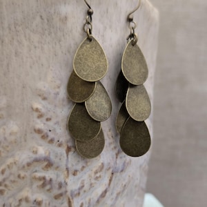May include: A pair of antique brass-colored earrings. Each earring features multiple teardrop-shaped pendants suspended from a hook. The earrings have a textured, aged appearance, suggesting a vintage or handcrafted style. The earrings are displayed against a neutral background.