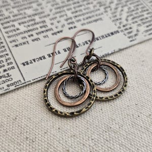 Puede incluir: Pendientes hechos a mano con tres círculos concéntricos en tonos bronce, cobre y plata. El círculo exterior tiene una textura martillada. Los pendientes cuelgan de ganchos para las orejas de color cobre, estilo bohemio.