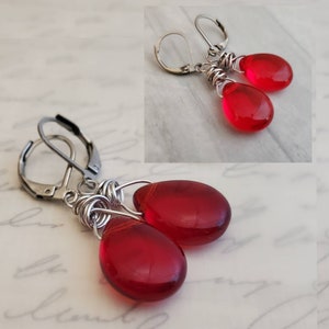 May include: A pair of red glass teardrop earrings with silver wire wrapped around the top of each teardrop. The earrings have silver leverback closures.