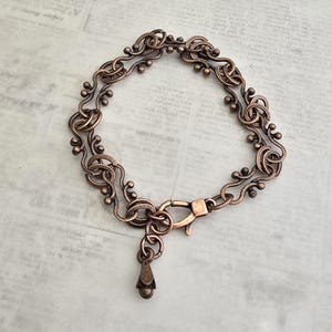 May include: A copper-colored bracelet with an intricate, flowing design. The bracelet features a series of interconnected loops and small, decorative beads. It has a lobster clasp closure and a small, teardrop-shaped charm hanging from the clasp.