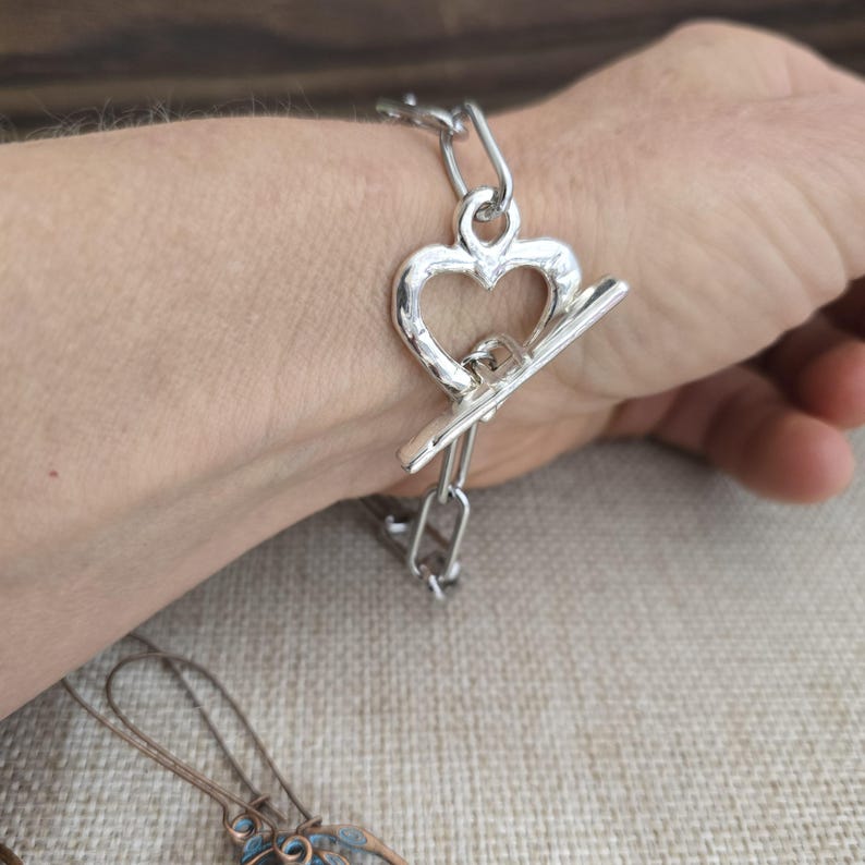 Pu&ograve; includere: Un braccialetto color argento con chiusura a levetta a forma di cuore. Il braccialetto &egrave; composto da segmenti di catena collegati. La chiusura a cuore &egrave; centrata sul polso. Il braccialetto &egrave; indossato al polso di una persona.