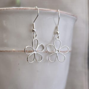 以下が含まれることがあります： 5枚の花びらを持つ花の形をしたシルバー調のドロップピアス。中央は透かし彫りになっており、シンプルなシルバー調のフックから吊り下げられています。清潔感のあるメタリックな仕上げと、左右対称の花の形が特徴です。