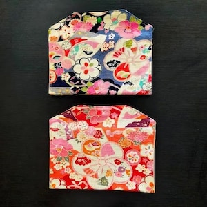 Op de afbeelding: Twee stoffen zakjes met een bloemenpatroon. Het bovenste zakje heeft een blauwe achtergrond met roze en witte bloemen. Het onderste zakje heeft een rode achtergrond met roze en oranje bloemen. Beide zakjes hebben een gebogen bovenrand.