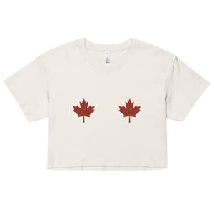 Op de afbeelding: Crèmekleurige crop top met twee rode esdoornbladontwerpen. De top met korte mouwen heeft een ronde halslijn en een relaxte pasvorm. Het esdoornbladontwerp is een symbool van Canada.