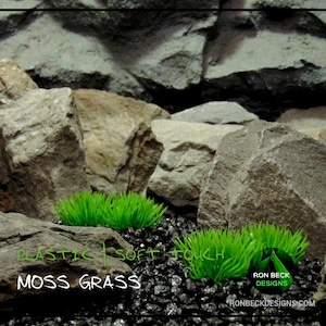 Puede incluir: Dos pequeños parches de hierba de musgo verde de plástico sobre una superficie de grava negra con grandes rocas grises en el fondo. El texto "Plastic | Soft Touch Moss Grass" está escrito en blanco en la parte inferior de la imagen.