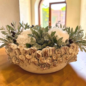May include: A round ceramic bowl with a textured, coral-like exterior featuring brown and cream tones. The bowl is filled with an arrangement of white flowers, green foliage, and small dark berries, creating a dense, layered botanical display.