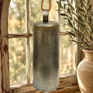 May include: A cylindrical metal bell with a weathered gray finish and gold-toned paint drips near the top rim. The bell features a sturdy metal loop at the center of the top, which is attached to a thick, natural-colored jute rope for hanging.