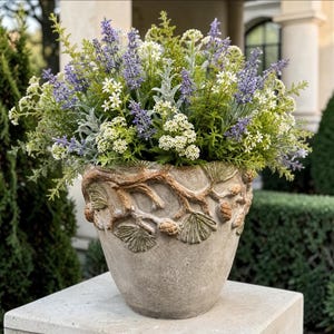 May include: A stone-colored planter featuring a raised relief design of pine branches and cones around the rim, filled with a dense arrangement of purple lavender sprigs, small white flowers, and various green foliage stems and leaves.