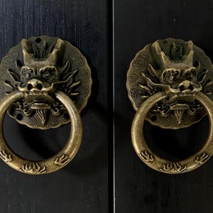 May include: Two antique brass-colored metal door knockers featuring a detailed dragon head design. Each circular backplate is embossed with coin motifs, and the swinging ring handle is decorated with small, stylized patterns in a metallic finish.