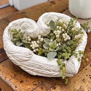 May include: A heart-shaped bowl with a textured, off-white finish resembling feathered wings. The bowl is filled with an arrangement of artificial greenery, including small round leaves, delicate white flowers, and clusters of tiny white berries.