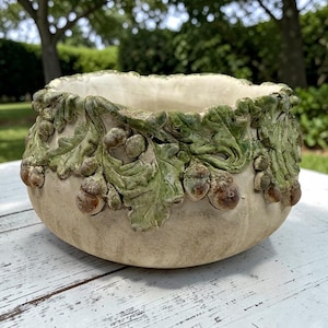 May include: A round ceramic bowl with a cream-colored base and a textured rim featuring sculpted green oak leaves and brown acorns. The interior of the bowl is smooth and off-white, while the exterior surface has a matte, weathered finish.