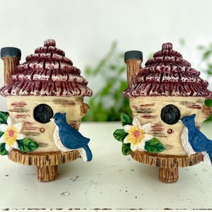 May include: Two identical decorative birdhouses featuring a textured cream-colored base, a dark red shingled roof, and a brown chimney. Each birdhouse has a circular entrance, a blue bird figurine on the side, and a white flower with green leaves.